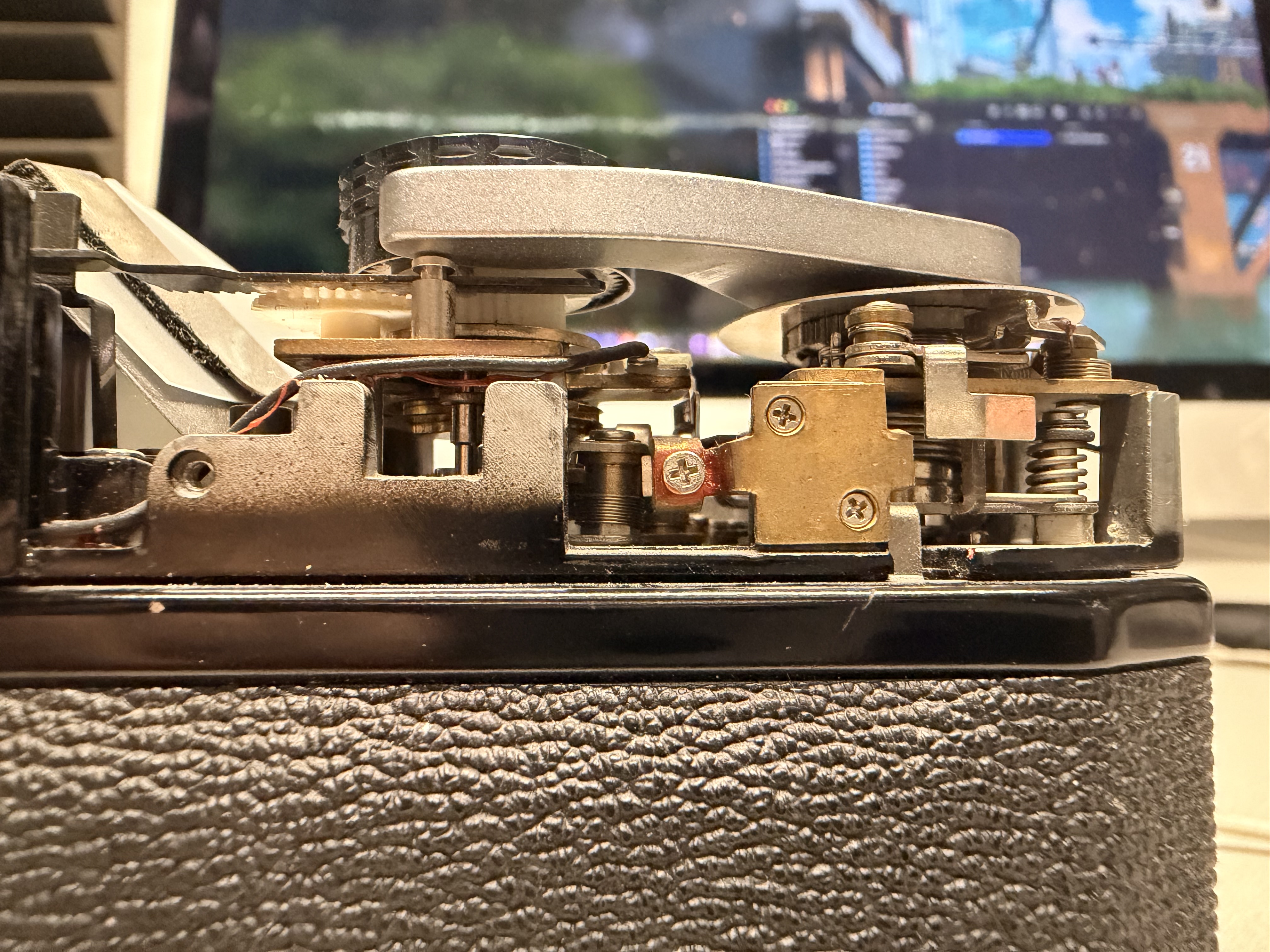 An extreme close-up, at eye level, of a film camera with its top plate lifted away. Exposed underneath are brass blocks, coiled steel springs, cross-head screws, a white nylon gear and the toothed rewind wheel, all seated on the camera's black leatherette body. A monitor glows out of focus behind it.